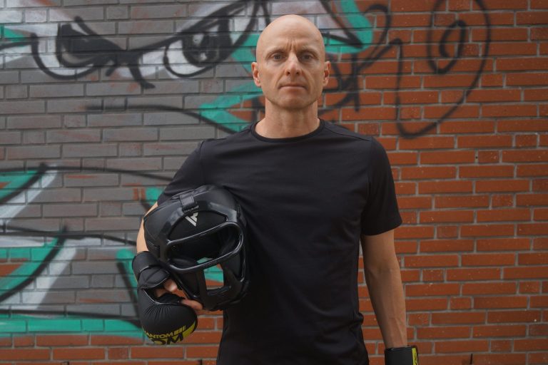 Selbstverteidigung Markus Koschorreck mit Helm mit Gesichtsschutz und MMA-Handschuhen vor Wand mit Graffiti.